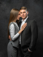 Man in business suit stands up straight and looks into camera. Next to him, girl gently presses herself against him. Studio portrait on dark background. Elegance, intimacy, style
