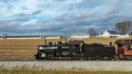 Fototapeta premium A classic steam train rolls along tracks beside vast farm fields under a cloudy sky, showcasing a nostalgic journey through the countryside. The train exudes an old-fashioned charm.
