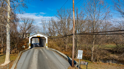 A historic covered bridge spans over a small rural road. The structure is framed by bare trees,...