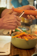 Close-up of a woman's hands peeling fresh carrots on a wooden countertop in a home kitchen. The image captures a moment of food preparation, highlighting home cooking, healthy eating, and rustic 