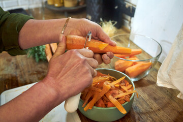 Close-up of a woman's hands peeling fresh carrots on a wooden countertop in a home kitchen. The image captures a moment of food preparation, highlighting home cooking, healthy eating, and rustic 