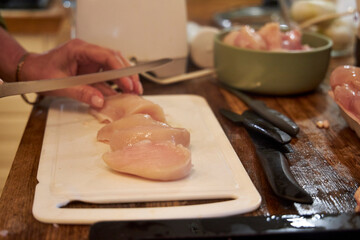 Close-up of a woman slicing raw chicken fillet into pieces on a white cutting board in a kitchen. The image captures a food preparation moment, focusing on healthy cooking, cleanliness, and domestic l