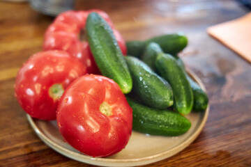 Close-up view of fresh washed tomatoes, cucumbers, and green onions arranged in a plate on a rustic wooden countertop. The image highlights healthy eating, fresh vegetables, and natural kitchen textur
