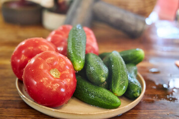Close-up view of fresh washed tomatoes, cucumbers, and green onions arranged in a plate on a rustic wooden countertop. The image highlights healthy eating, fresh vegetables, and natural kitchen textur