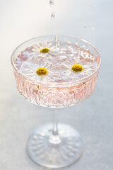 Elegant crystal glass filled with refreshing pink beverage garnished with floating daisies. Droplets splashing in the glass, creating a fresh, summery atmosphere against a soft light background.