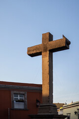 Cruz de piedra gris en el atardecer. Paloma postrada sobre una cruz de concreto iluminada por la luz del sol.  De fondo un cielo limpio y azul.