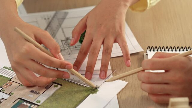 Architects using measurement tool and pencil tracing in building blueprint by coworker support on table with architectural document and wooden block scatter around at office. Closeup. Delineation.