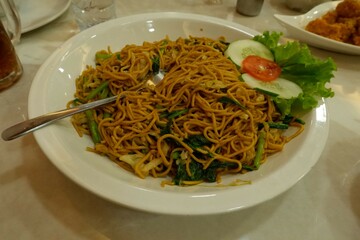 A white plate filled stir fried of cooked noodles with green vegetables fresh lettuce, cucumber, broccoli and tomato slice garnished by pieces of chicken, egg or tofu. fork is placed on top the noodle