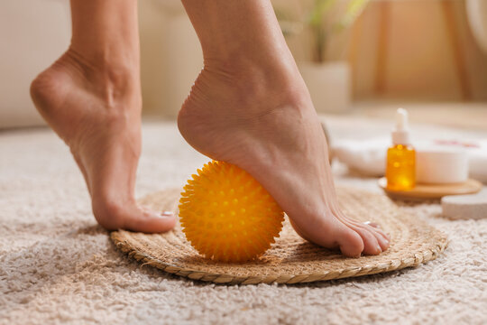 Close-up of a woman's foot rolling on a yellow spiky massage ball during a foot care routine at home with spa oils and cream in soft natural light. Therapy and wellness concept.