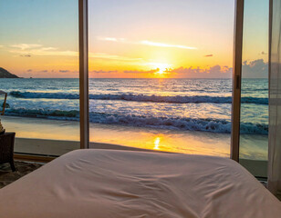 A cozy bed faces a breathtaking ocean view through large glass windows, as the golden sun sets over the horizon, casting warm reflections on the gentle waves.