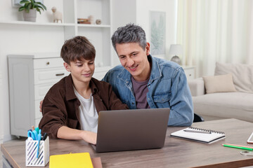 Smiling father and his son doing homework with laptop at table indoors