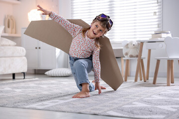Cute little girl with cardboard plane wings and goggles playing pilot at home