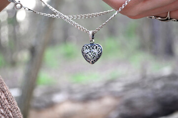 Fototapeta premium an elegant silver pendant in the shape of a heart with an openwork pattern, suspended on a chain. image of nature - greenery and light tree trunks. The pendant is held by the hand, creating an accent