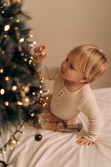 Blond toddler in warm home clothes curiously explores Christmas tree decorations reaching for golden ornament with fascination and wonder in cozy festive atmosphere Little child reaches for golden bal