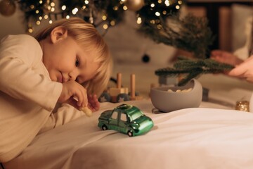Blond toddler lies near glowing Christmas tree playing with green toy car holding puffed snack in warm festive bedroom full of love care joy and everyday moments of modern motherhood