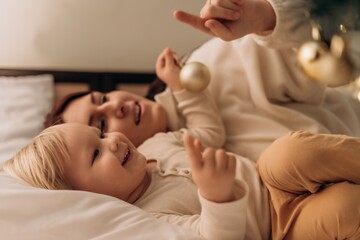 Smiling toddler lies in bed beside mother pointing at Christmas tree lights while holding ornament sharing magical moment of love togetherness warmth joy and connection in modern motherhood