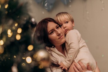 Mother holding child near glowing Christmas tree as both gaze forward wrapped in gentle emotion soft light and peaceful atmosphere expressing tenderness love and togetherness of modern motherhood