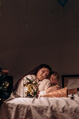 Loving mother gently embraces her sleepy child under warm lights as peaceful festive evening fills the room with comfort closeness and the magic of shared tenderness A soft tender moment where mother