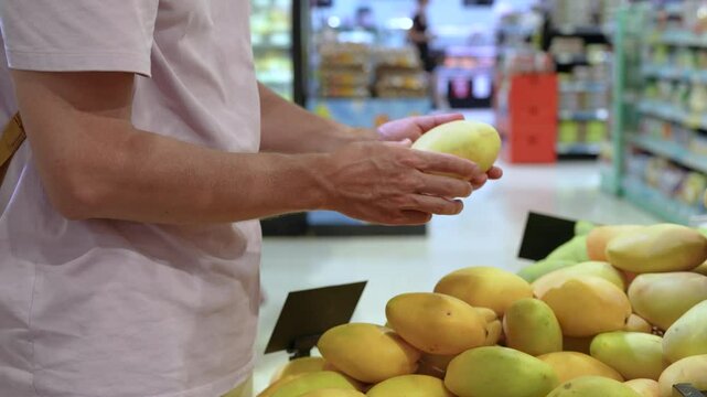Unrecognizable man examing mango fruit, squeezing to test ripeness before buy. Fresh tropical fruit selection, grocery shopping, travel destination in grocery market, local produce quality checked