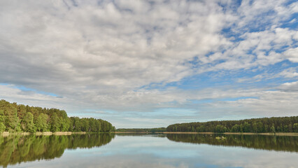 Sunny summer landscape on the lake. Reflection of the sky and clouds in the water. A valley of green trees on the shore of a lake. A concept for travel and camping.