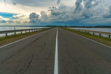 An empty asphalt road runs through the river. Thick gray clouds over the highway along the lake.