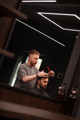 Barber trims a client's hair at a modern barbershop, seen through a mirror. The focused barber uses...