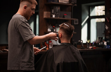 Barber gives a precise haircut to a seated male client using clippers in a stylish, modern barbershop. Shelves with grooming products and sunlight from the window enhance the cozy workspace..