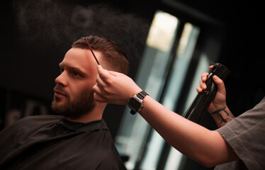 Fototapeta premium Barber sprays mist on a client's styled hair while combing the front in a well-lit modern barbershop. The client sits calmly, showcasing a fresh, sharp look and professional grooming care..