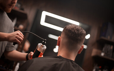 A male barber sprays liquid on a client's freshly cut hair in a modern barbershop. The client sits with his back to the camera, showcasing a clean fade haircut under stylish lighting..