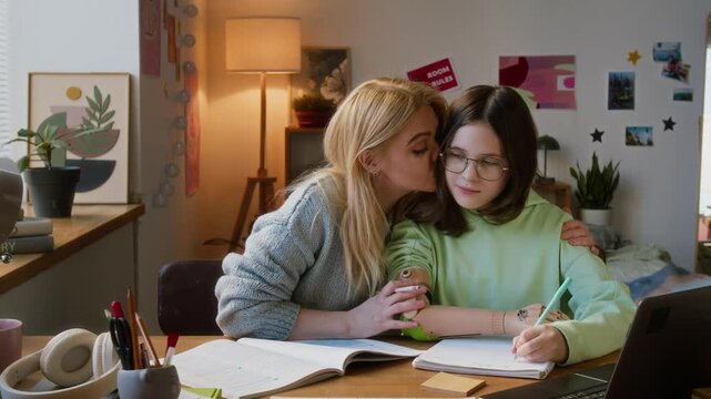 Loving mother hugging and kissing on cheek her teenage daughter with prosthetic arm while helping her do her homework
