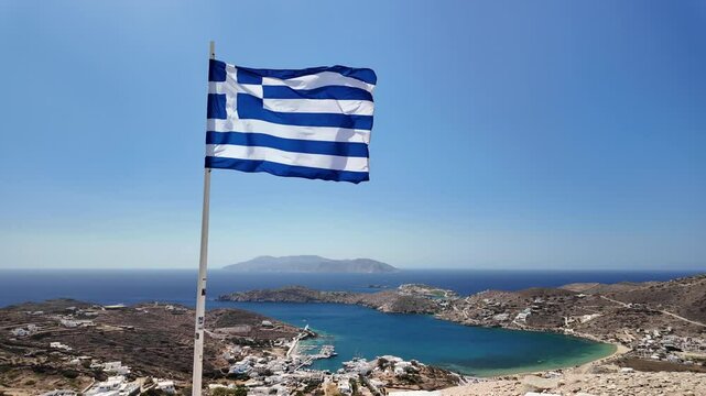 View of the Greek national flag overlooking the picturesque port of Ios Greece and the island of Sikinos in the background in slow motion
