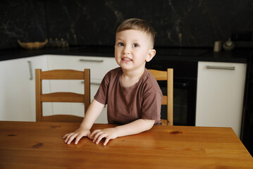 Naklejka premium Cute little boy in a brown T-shirt sitting at a wooden table, smiling and looking slightly upward with a cheerful expression.