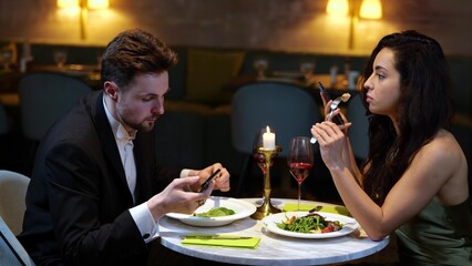 Smiling couple having date in the restaurant while eating salads and holding smartphones....