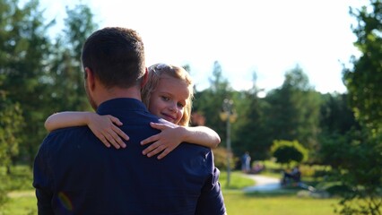 Back view of man walking in nature holding his little daughter in arms. Pretty child girl smiling to camera hugging father outdoor in park. Summer weekend. Family day. Childhood concept