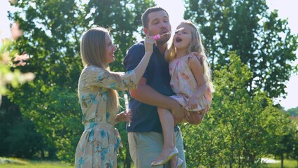 Joyful family spending summer day together outdoor playing with soap bubbles. Parents play with child on a walk. Girl blowing bubbles with father and mother in park. Childhood concept