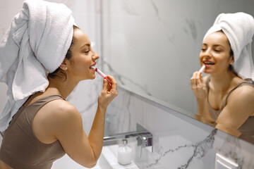 Pretty young woman with towel on her head puting lipstick on her lips looking in the mirror near...