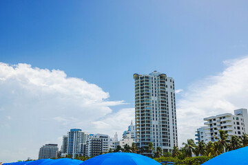 Modern coastal skyscrapers in Miami Beach against blue sky with palm trees and blue umbrellas on Atlantic Ocean shore.