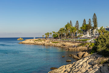 Coastal resort in Cyprus with palm trees, rocky shore, and clear blue sea on a sunny day.