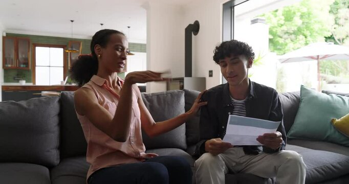 Diverse mother and son opening envelope on couch, reading printed letter, celebrating good news