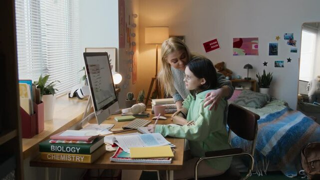 Caring mother checking up on her teenage daughter with prosthetic arm doing her homework at desk in her cozy bedroom