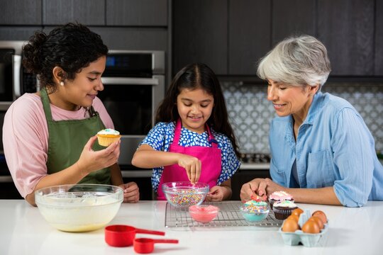 Three generations of women happily decorating cupcakes together in a modern kitchen with colorful sprinkles and frosting bowls