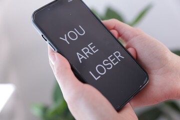 Cyber bullying. Woman holding smartphone with text You Are Loser on blurred background, closeup