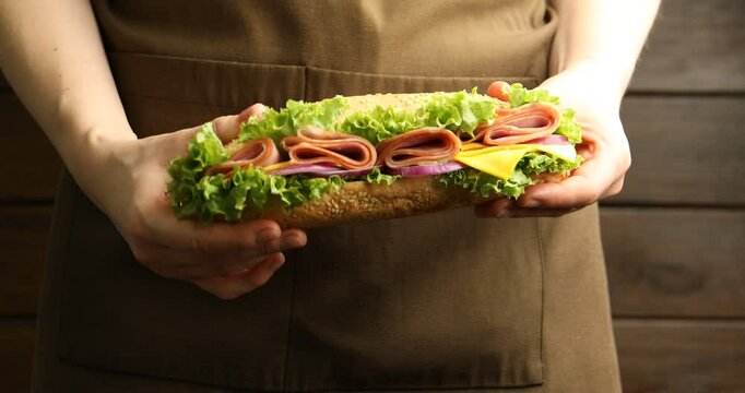 Woman holding tasty baguette sandwich with ham, cheese and vegetables near wooden wall, closeup
