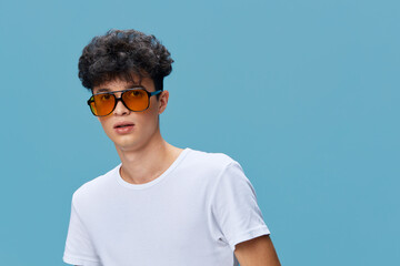 young man wearing white t-shirt and orange sunglasses posing against a blue background with curly hair and casual style, modern and trendy look