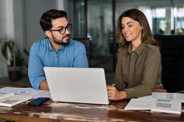 Two smiling busy professional business people working in office with laptop computer. Female executive manager talking to male colleague or partner having conversation sitting at workplace.