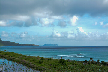 A beautiful view of the turquoise ocean meeting the shore, with lush green vegetation along the coast and distant islands under a partly cloudy sky.