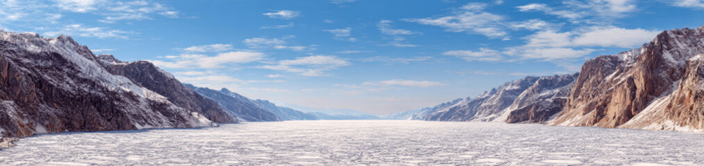 Frozen mountain valley with icy lake under bright blue sky and scattered clouds, creating serene and cold winter landscape