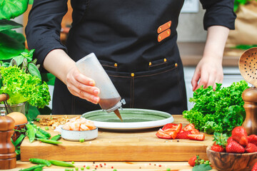 Chef cooking food in the kitchen. Cook drips sauce onto plate prepares gourmet lettuce salad with strawberries and shrimp, wooden table with kitchenware, vegetables, herbs and ingredients. 