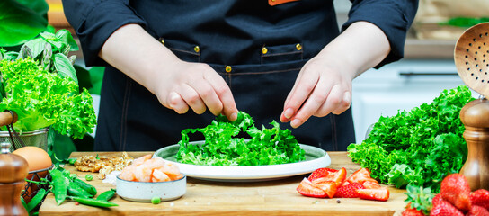 Chef cooking food in the kitchen. Cook prepares gourmet lettuce salad with strawberries and shrimp., wooden table with kitchenware, vegetables, herbs and ingredients. 