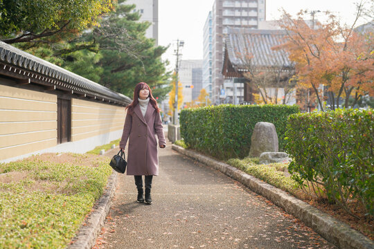 On a chilly December morning, a Japanese woman in her early twenties with long pink hair and a pink trench coat walks a quiet street lined with wooden houses, embracing winter's gentle calm.